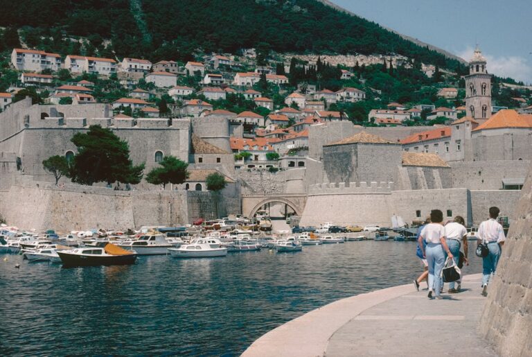 10 Essential Travel Tips for Visiting Dubrovnik โ Travel Guide to Dubrovnik 158 People walk along a harbor with boats and walled city.